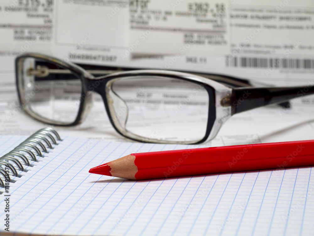 red pencil on a spiral notebook with reading glasses and bills in background red pencil on a spiral notebook with reading glasses and bills in background