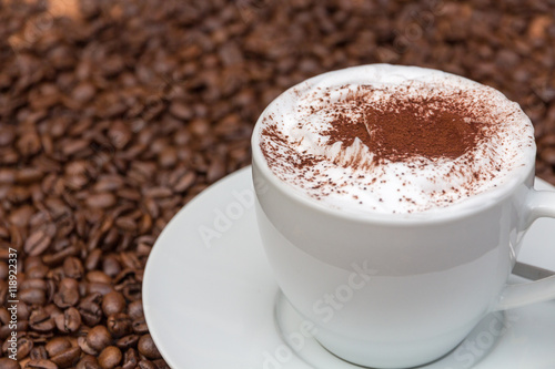Cappuccino trinken, weiße Kaffeetasse mit Milchschaum und Kakaopulver stehen auf Kaffeebohnen