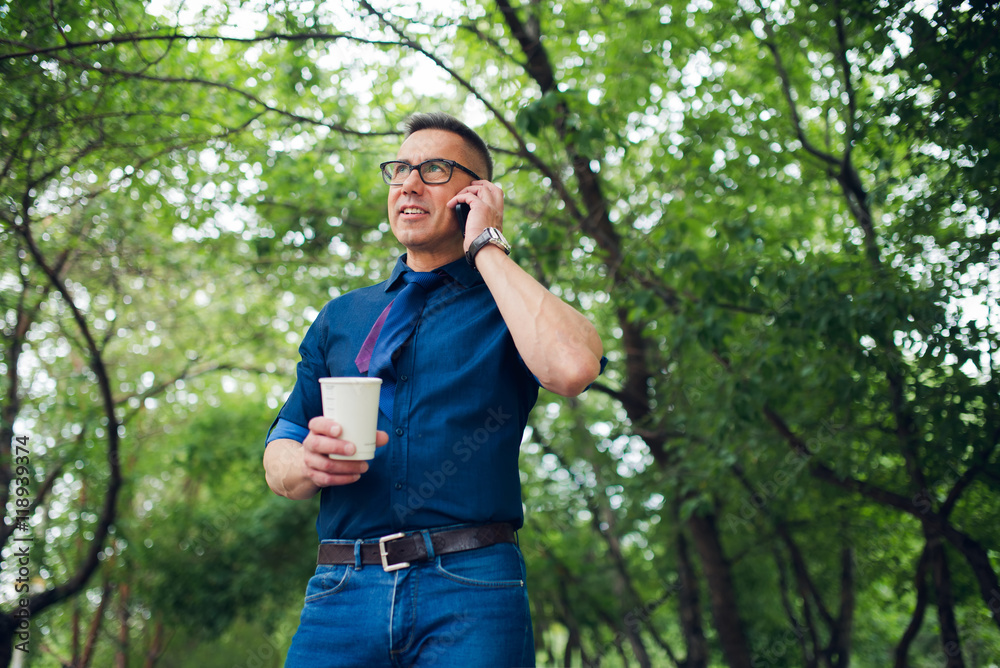Mature man talking on his cellphone while standing with coffee in park