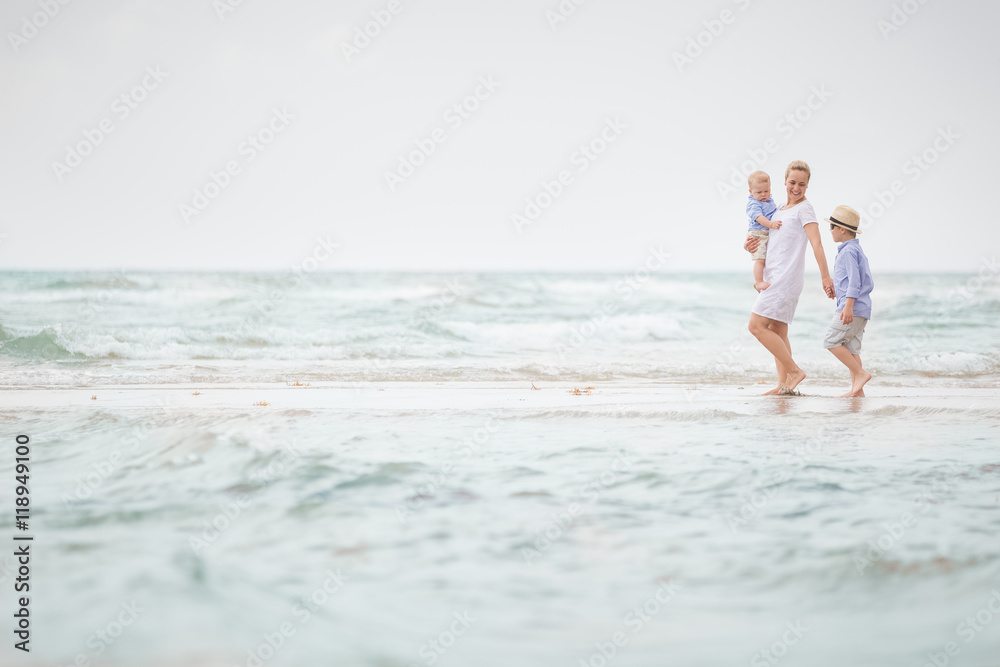 Young mother in white dress walking with her two little boys along the ocean beach. Woman with baby and boy enjoying vacation by the sea. On the empty beach. Motherhood. Water background. copy space.
