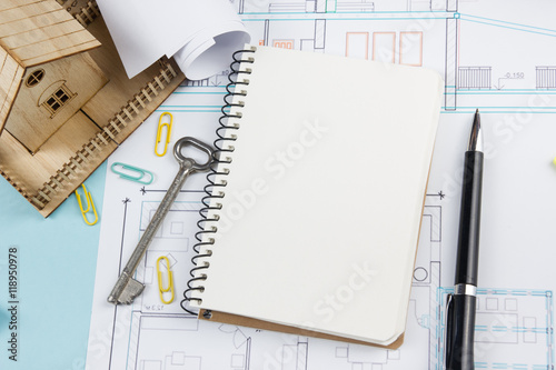 Real estate concept. Blank white notebook on architectural desk table blueprint background with key, pen, small house, office supplies. Copy space for ad text, top view.