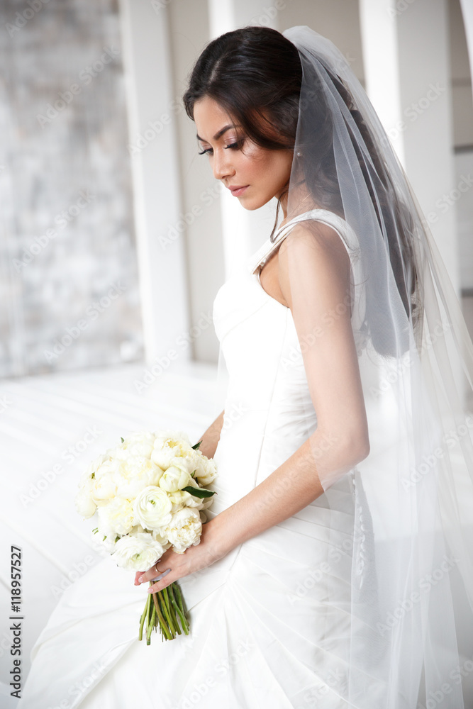 Portrait of a beautiful and embarrassment slim bride with the bouquet of peonies in the elegant classic white wedding dress with long veil. Light studio shot Portrait of a beautiful and embarrassment slim bride with the bouquet of peonies in the elegant classic white wedding dress with long veil. Light studio shot
