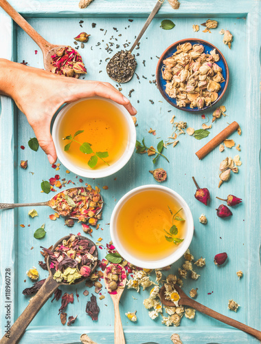 Two cups of healthy herbal tea with mint, cinnamon, dried rose and camomile flowers in spoons, human's hand taking cup, blue background, top view