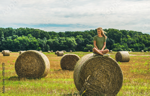 Fototapeta Young blond lady sitting legs crossed on haystack and smiling on sunny summer da