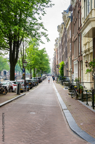 Narrow Cobbled Street lined with Trees and Brick Building alongside in Amsterdam © alpegor