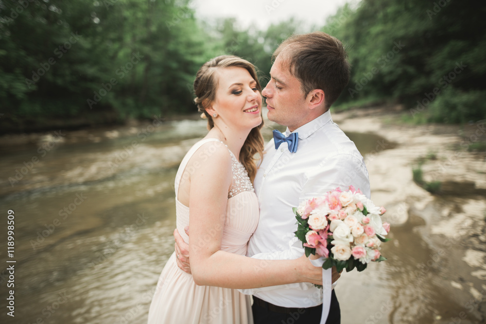 Beautifull wedding couple kissing and embracing near the shore of a mountain river with stones Beautifull wedding couple kissing and embracing near the shore of a mountain river with stones