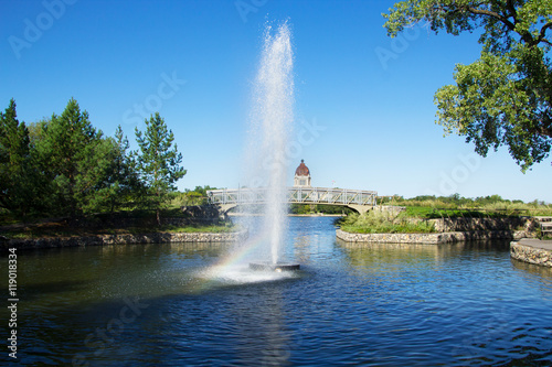Early morning photo of a fountain in Regina Saskatchewan's Wascana Park with a rainbow in the spray. Landscape view with a footbridge and the provincial legislature in the distance. © hihatimages