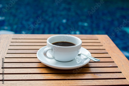 Cup of black coffee set on wooden table at blue pool background. Coffee by the pool in the morning
