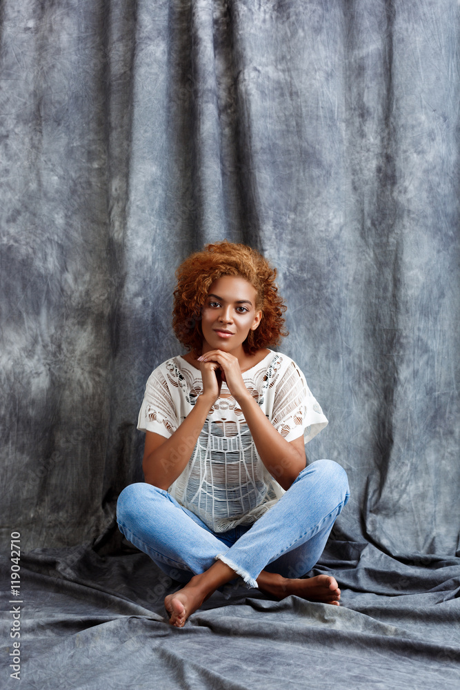 Fototapeta premium Young beautiful african girl sitting at floor over grey background.