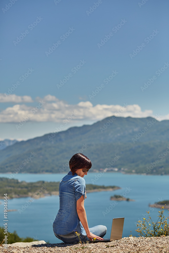 Fototapeta premium Beautiful young freelancer woman using laptop sitting on the lake.Happy smiling girl working online.Studying and learning using notebook computer.Freelance work,business people concept.