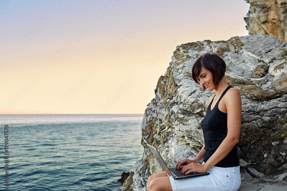 Beautiful young freelancer woman using laptop sitting near the sea.Happy smiling girl working online.Studying and learning using notebook computer.Freelance work,business people concept.