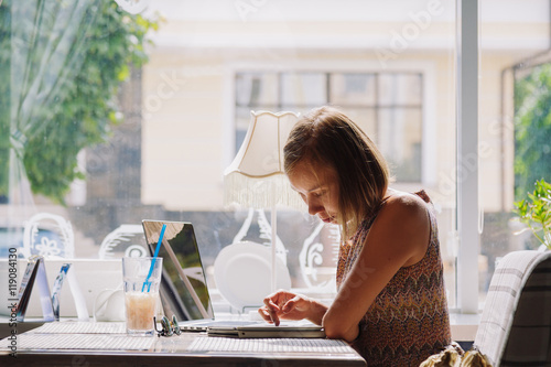 Side view of young short-haired woman in dress using laptop while sitting at table with drink and tablet in cafe against of window