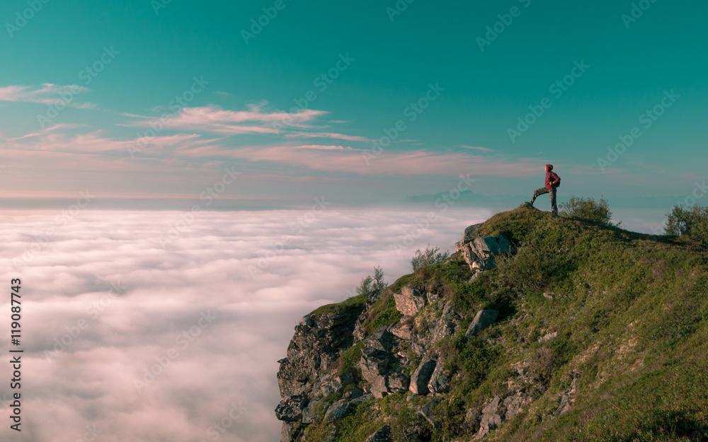 Toned image adult woman with a backpack stands on the edge of a cliff and looking at the sunrise against the blue sky and thick clouds floating down