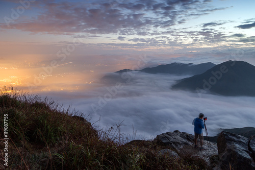 Man photographing the lights of Tung Chung New Town below clouds on Lantau Island. Viewed from the Lantau Peak (the 2nd highest peak in Hong Kong, China) at dawn.