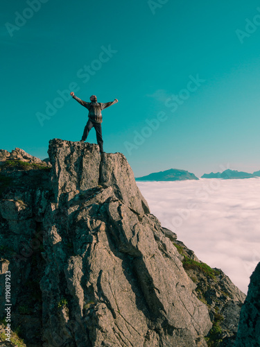 Toned image adult man with backpack stands on the edge of a cliff and screams and shows his hand into the distance against the blue sky and thick clouds floating down