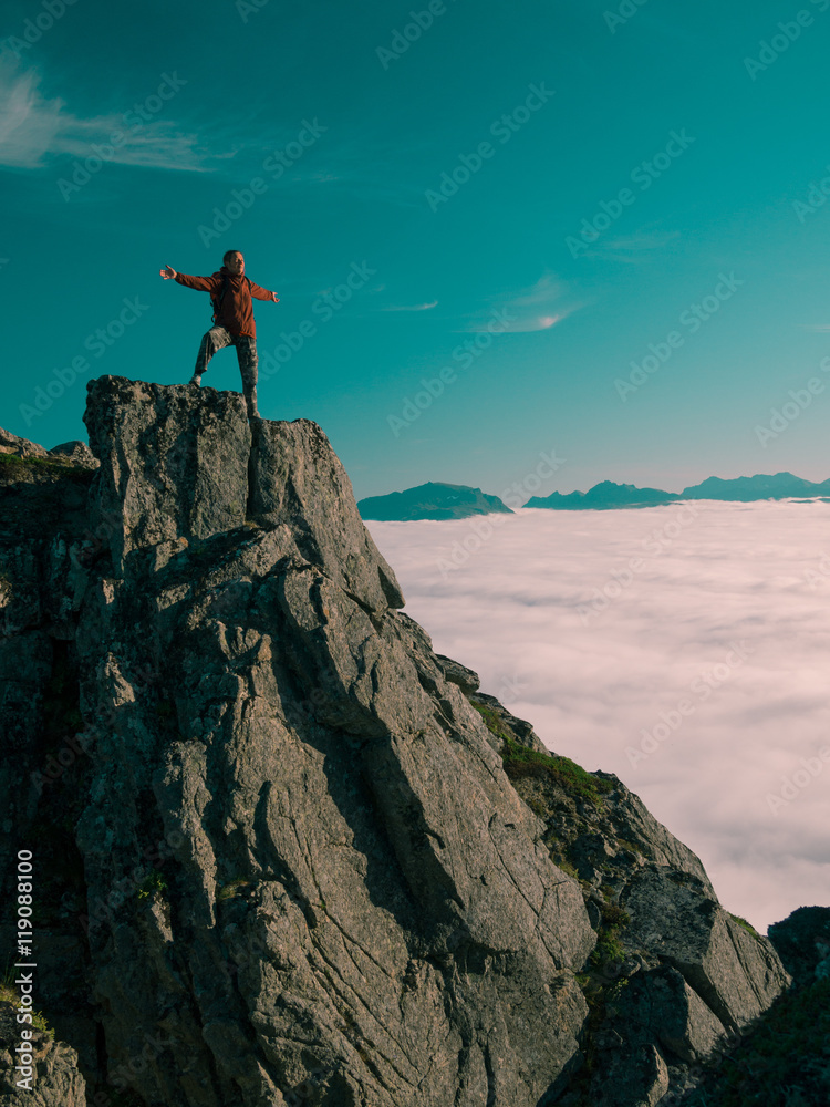 Toned image adult woman with a backpack with arms outstretched stands on the edge of a cliff and looking at the sunrise against the blue sky and thick clouds floating down