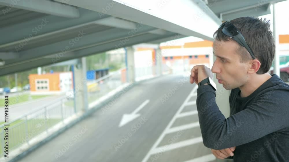 Outdoor portrait of modern young man with smart watch in the street. The man in glasses with backpack sitting on the bridge. Bottom drive cars Outdoor portrait of modern young man with smart watch in the street. The man in glasses with backpack sitting on the bridge. Bottom drive cars