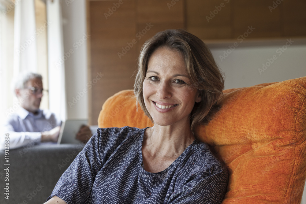 Smiling woman relaxing in armchair with husband in background Smiling woman relaxing in armchair with husband in background