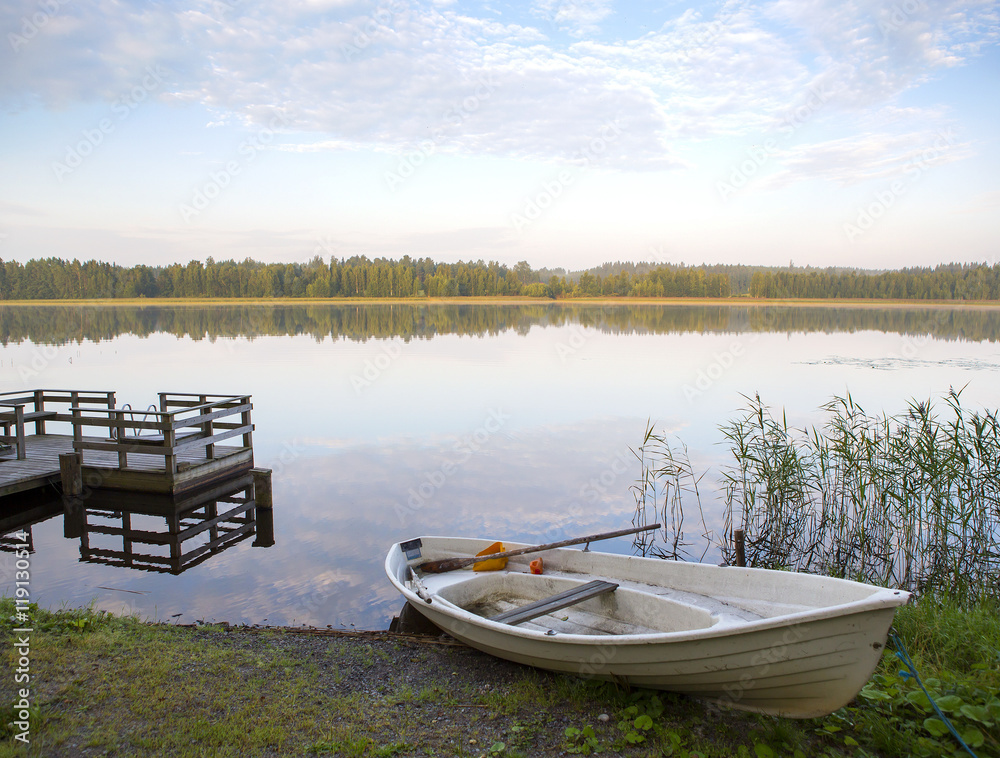 Silent morning. A rowing boat is waiting next to an empty pier in the morning sun. The sun is rising from the backside. Still water reflects the forest.