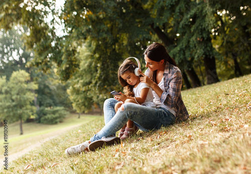 Mother and daughter sitting on grass and listening to music on smart phone.