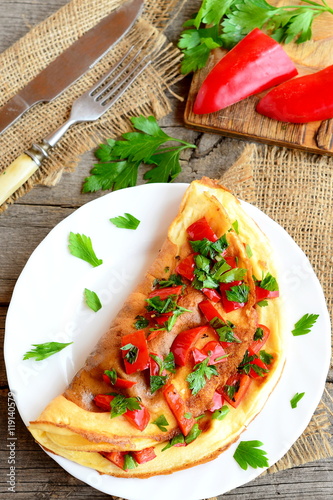 Stuffed egg omelette with vegetables on a plate. Healthy omelet with fried red pepper, fresh parsley and spices. Fork, knife on a burlap and old wood background. Easy eggs recipe. Top view