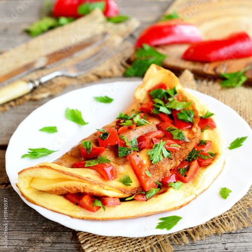 Fried omelette with vegetables on a plate, fork and knife on old wooden background. Colorful omelette stuffed with roasted red pepper and chopped fresh parsley. Breakfast recipe with eggs. Closeup