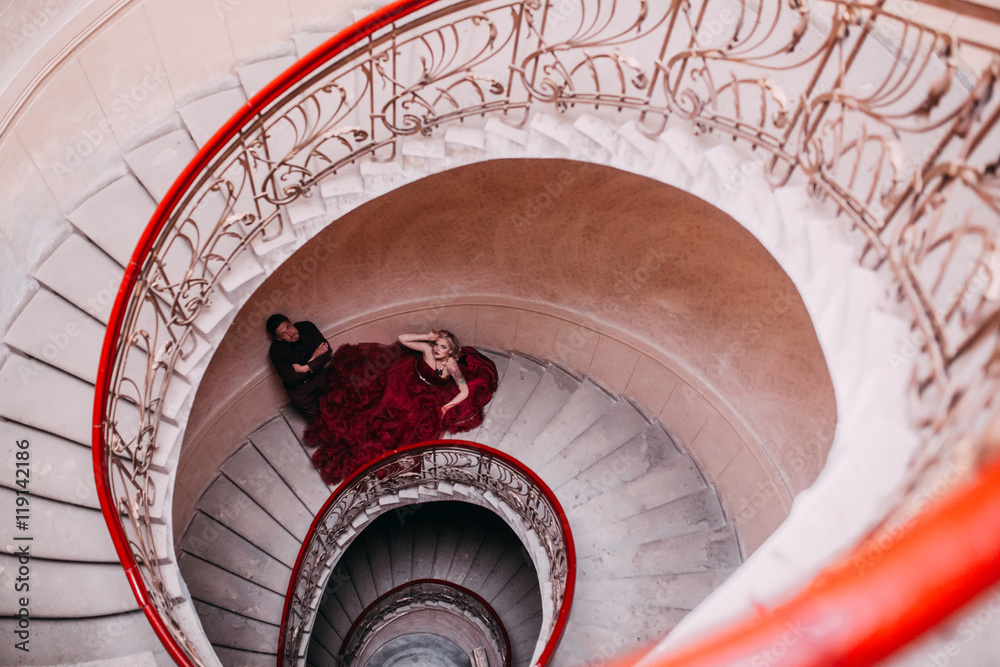 Elegant couple at old vintage house and palace with big round stairs. Man in brown shirt. Woman in red cloud dress. 