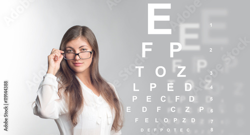 Молодая красивая девушка в очках, серый фон с таблицей для проверки зрения 
