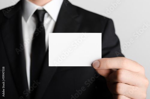 Business Card Mock-Up (85x55mm) - Man in a black suit holding a blank card on a gray background.