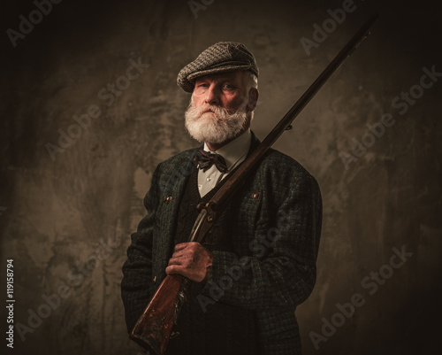 Senior hunter with a shotgun in a traditional shooting clothing, posing on a dark background Fototapet