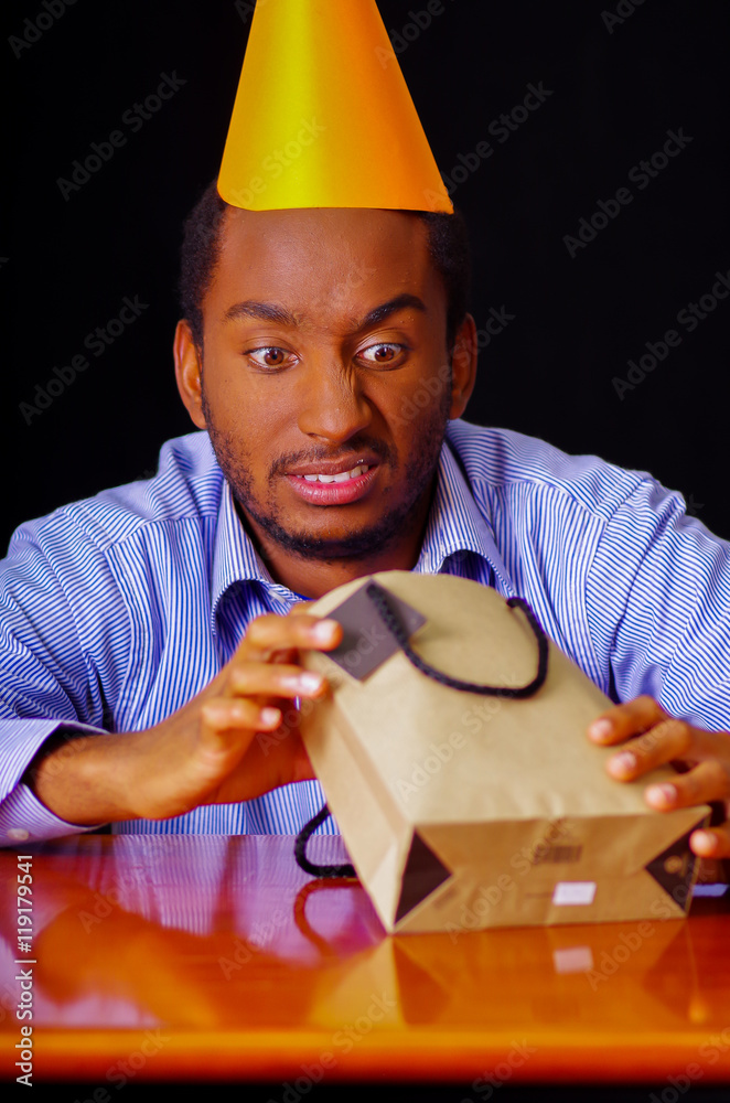 Handsome man wearing blue shirt and hat sitting by table opening birthday present, looking upset, celebrating alone concept Handsome man wearing blue shirt and hat sitting by table opening birthday present, looking upset, celebrating alone concept