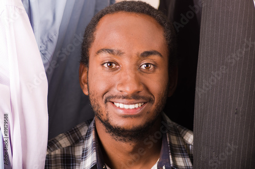 Headshot handsome young man standing inside wardrobe with clothes sorrounding, smiling to camera