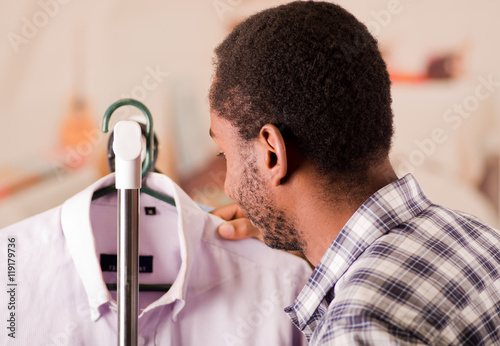 Handsome young man standing inside wardrobe going through rack of different shirts hanging, fashion concept