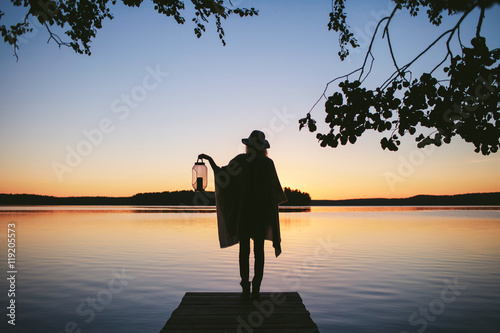 Fotografia girl in a poncho and a hat with lamp standing back to the view of the sunset on