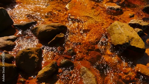 Stream water full of red ferric sediments. Cascades in stream, mossy boulders and terrible smell red mud. Bubbles of gas, stalk of fern and grass. Obscene destruction of the environment and nature