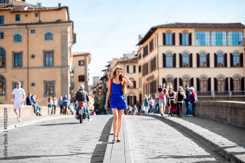 Beautiful female traveler in blue dress, hat and backpack walking on crowded Holy Trinity bridge in Florence city. Vacation in Italy