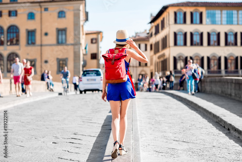 Beautiful female traveler in blue dress, hat and backpack walking on crowded Holy Trinity bridge in Florence city. Back view