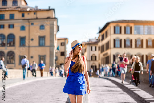 Beautiful female traveler in blue dress, hat and backpack walking on crowded Holy Trinity bridge in Florence city. Vacation in Italy
