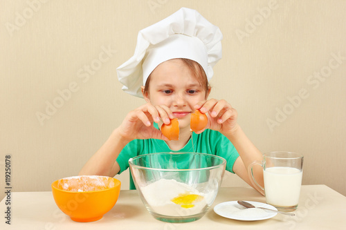 Young smiling boy in chefs hat pours eggs for baking the cake