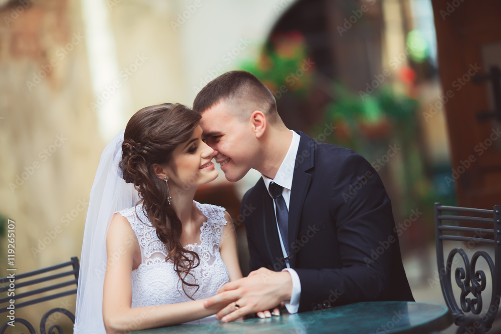 Wedding couple in love. Beautiful bride in white dress and veil and brides bouquet with handsome groom in blue suit sitting in cafe. Full lenght portrait of man and girl. Concept of wedding