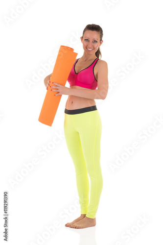 Smiling brunette woman in sports neon yellow leggings and pink bra standing with the orange rolled mat after training on white background