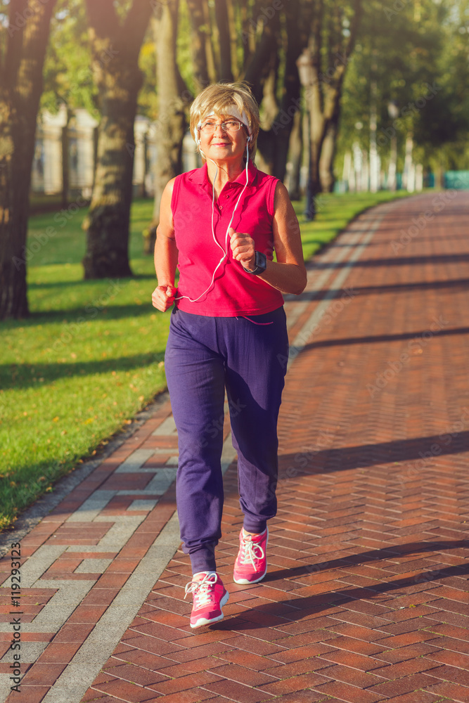 Elderly woman jogging with headphones and smartwatches in the park in evening sunset Elderly woman jogging with headphones and smartwatches in the park in evening sunset