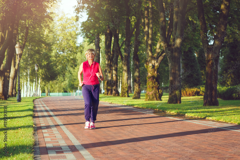 Elderly woman running with headphones and smartwatches in the park in evening sunset Elderly woman running with headphones and smartwatches in the park in evening sunset