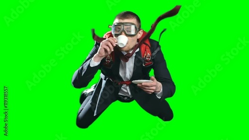 Calm and relaxed young businessman flying in free fall drinking coffee, wearing formal suit, aviator mask and parachute on his back, chroma key against green screen background