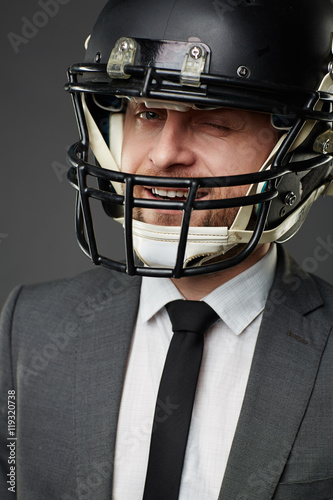 Portrait of smiling businessman wearing suit and American football helmet winking at camera, isolated on black background.