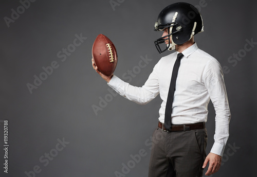 Sideview of businessman in formal clothes and football helmet standing isolated on black background looking in determination at American football. photo