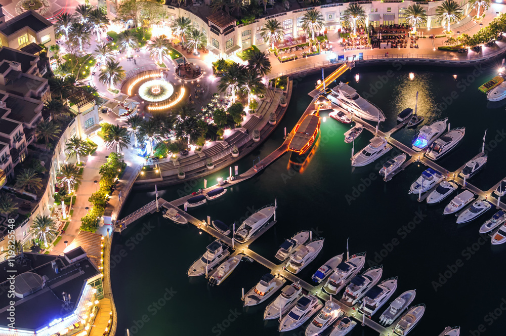 Fototapeta premium Amazing colorful dubai marina fountain and yacht dock during night. Beautiful sidewalk among tallest skyscrapers of the world. Dubai marina, United Arab Emirates.