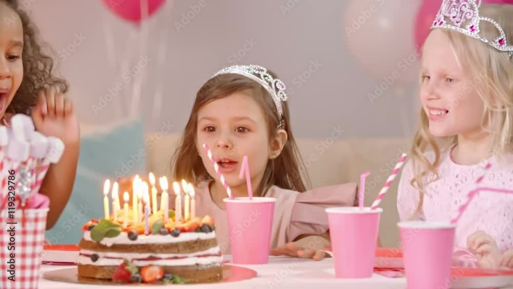 Little girls with tiaras on their heads taking off hands from face and getting surprised about birthday cake on table. They blowing out candles together and clapping hands