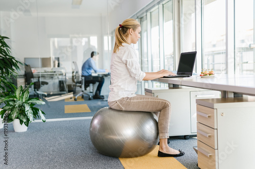 Business woman working on laptop in office while sitting on pilates ball photo