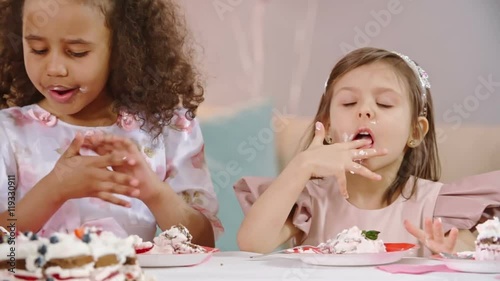 Adorable little princesses eating birthday cake at the table and licking fingers in slow motion photo